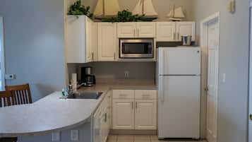 A compact kitchen with white cabinets, a microwave, a refrigerator, and a sink. There are two sailboats on top of the cabinets.