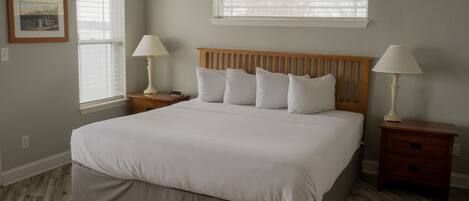 A bedroom with a large bed, two bedside tables with lamps, a window with blinds, and a framed picture on the wall.