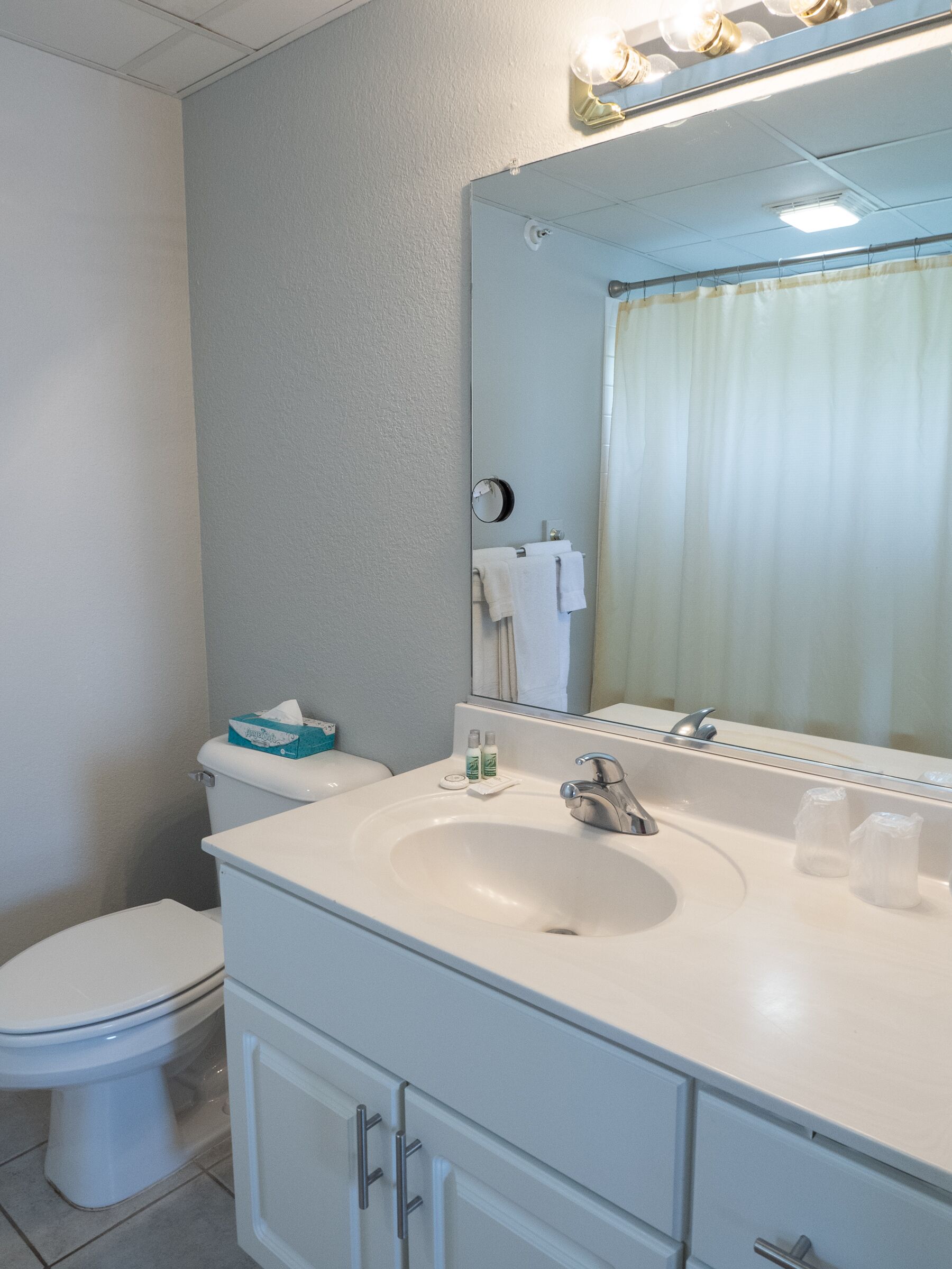 A bathroom with a white sink, mirror, and toilet. There are toiletries on the sink and a towel rack above the toilet.