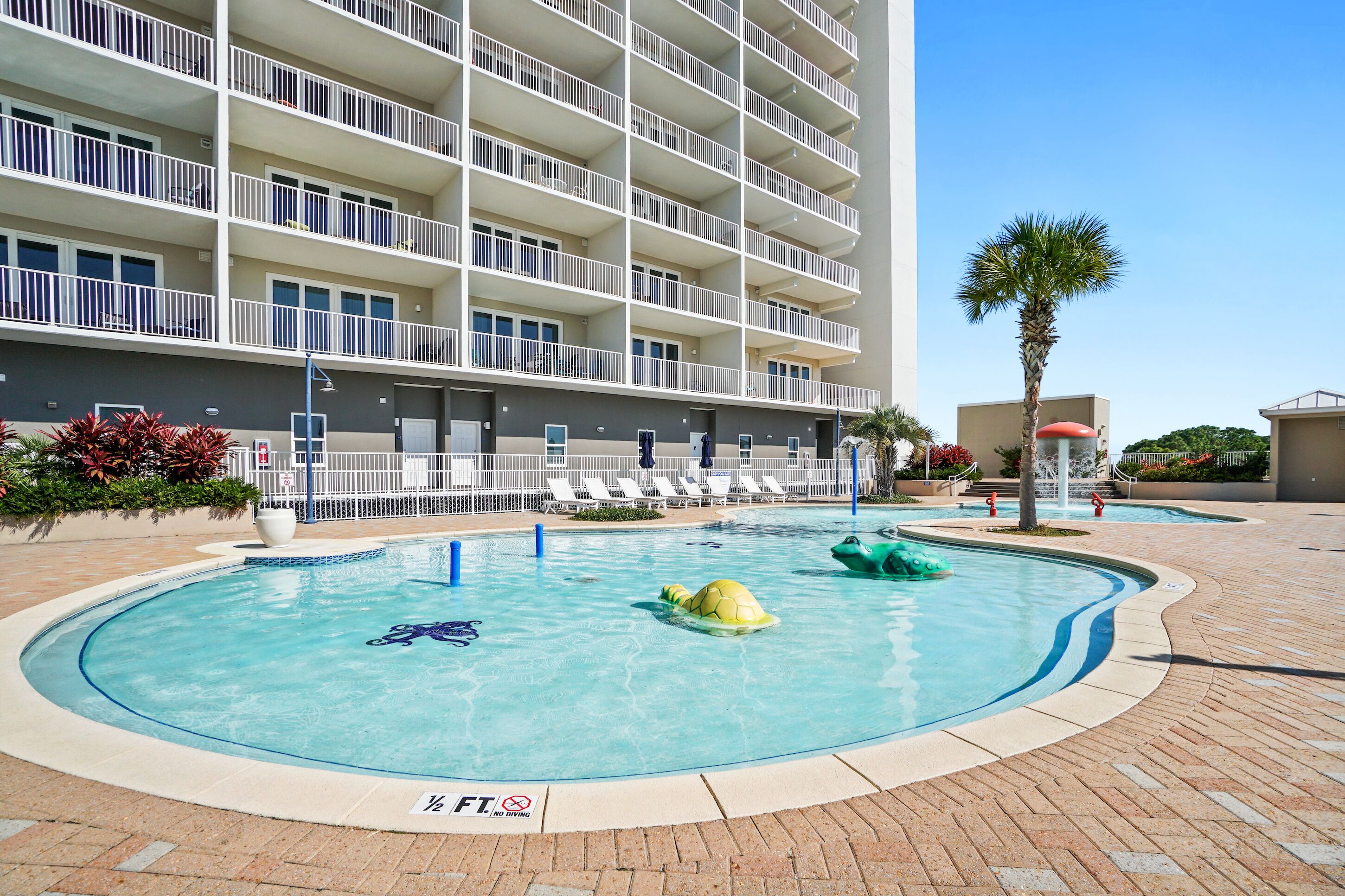 A swimming pool with lounge chairs, a palm tree, and a multi-story building with balconies.