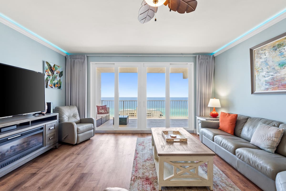 A living room with a TV, leather sofa, and a view of the beach.