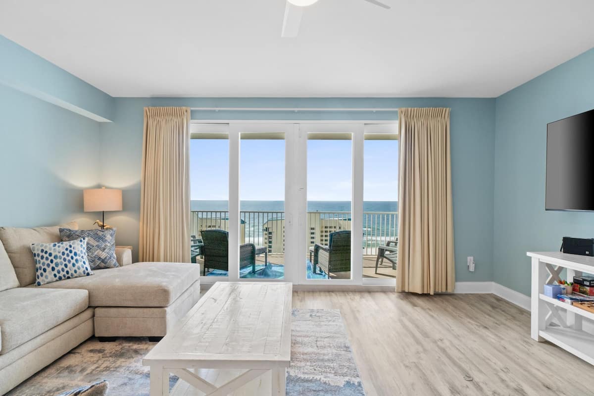 A living room with a sofa, coffee table, and a TV. There is a balcony with outdoor furniture.