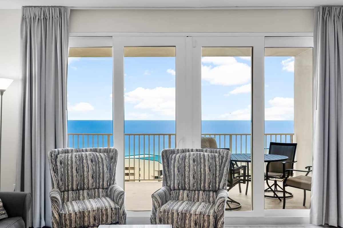 A room with two patterned armchairs, a table, and a view of the ocean through large sliding glass doors.