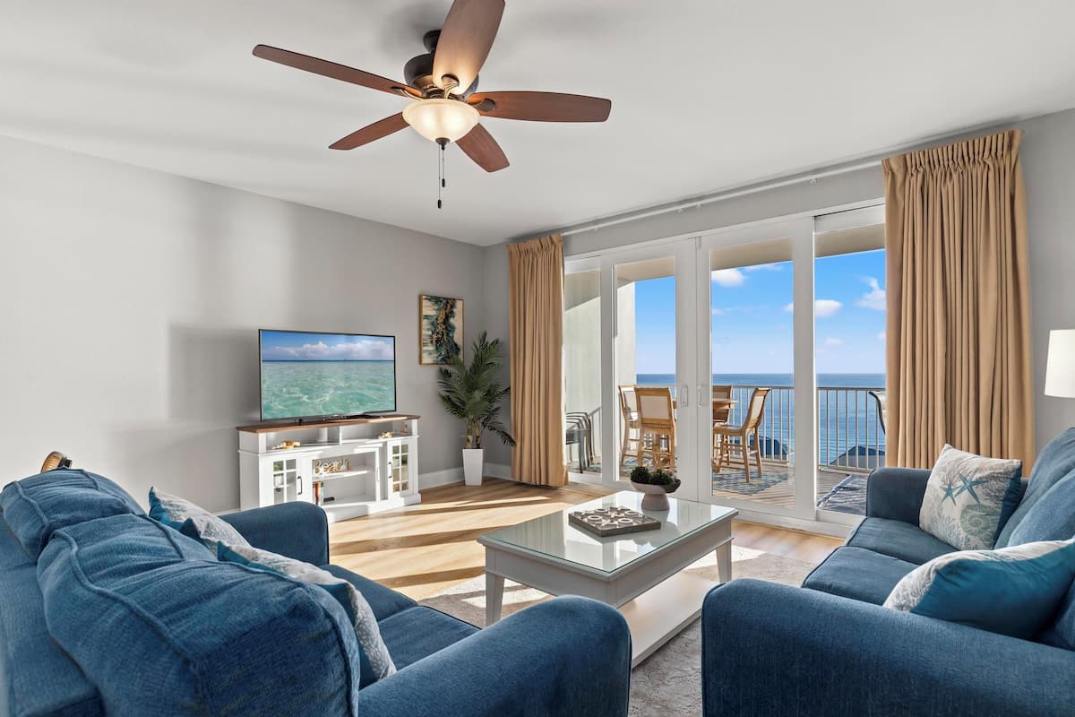 A spacious living room with a ceiling fan, a flat-screen TV, and a balcony with a view of the ocean.
