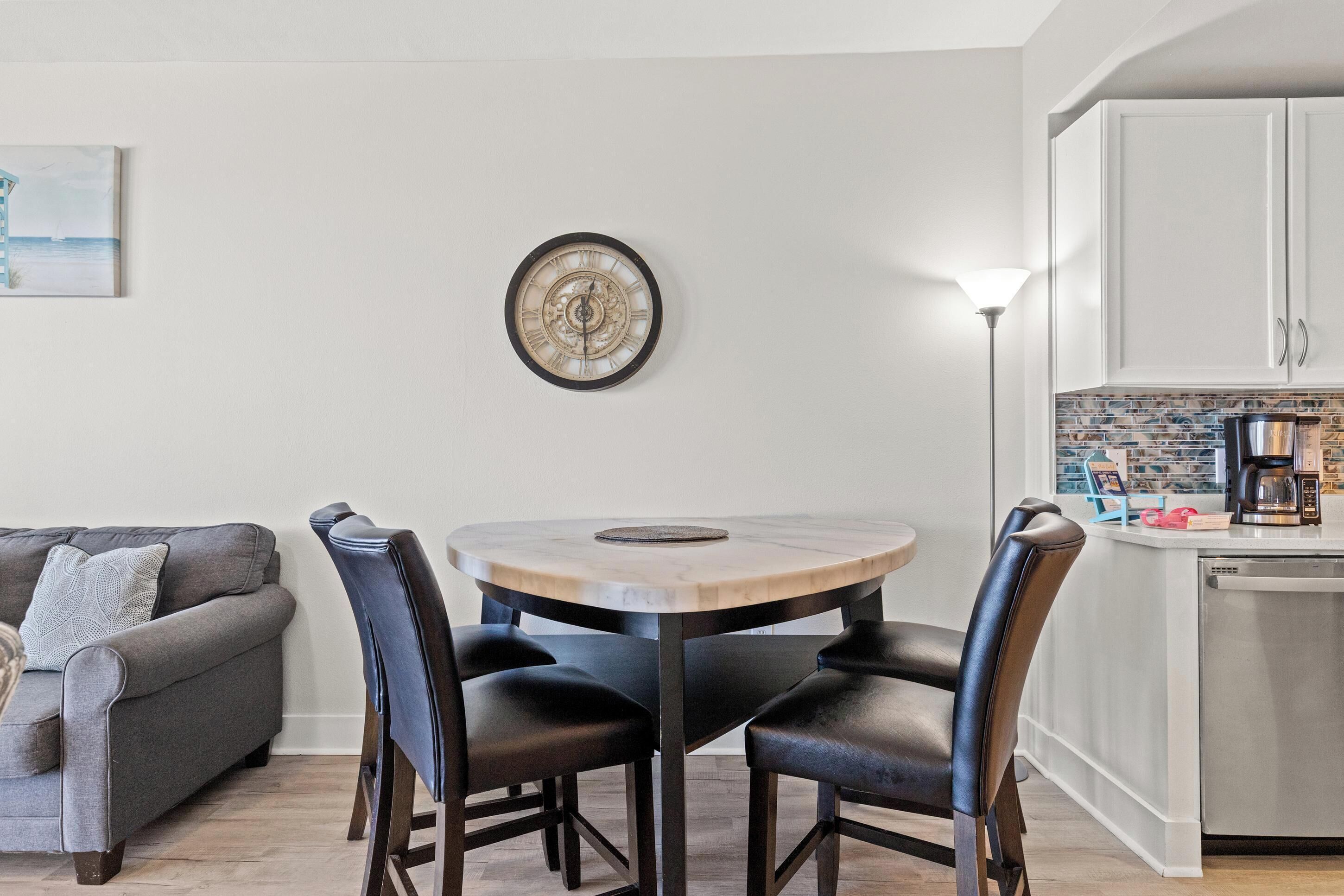 Comedor con una mesa redonda, cuatro sillas, un reloj de pared, un cuadro, un sofá y una zona de cocina con armarios y electrodomésticos.