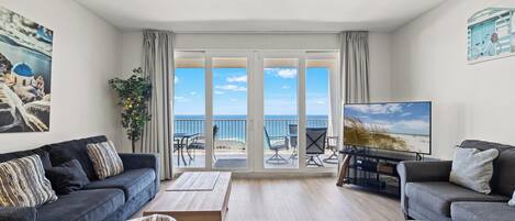 A living room with a sofa, a coffee table, and a TV. There is a balcony with a view of the sea.