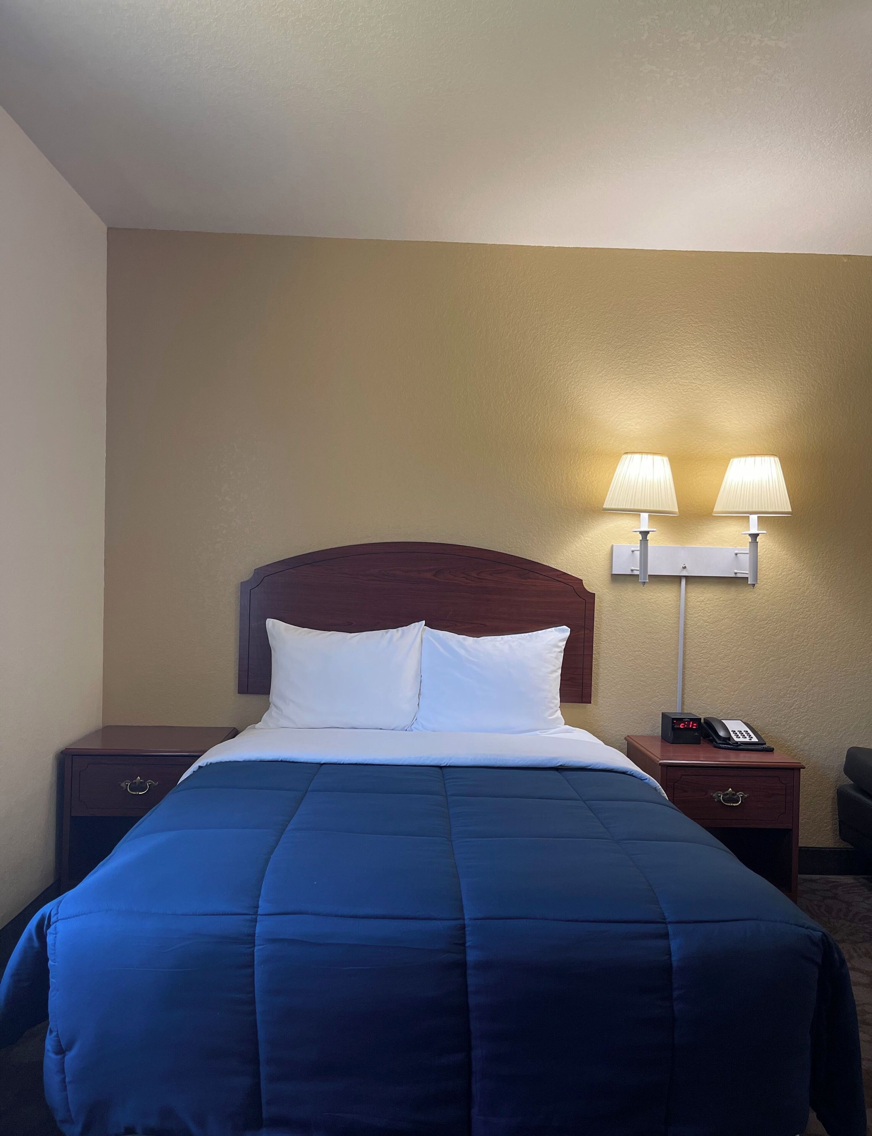 Une chambre d’hôtel avec un lit, deux lampes de chevet, une table de chevet avec un téléphone et une horloge murale.