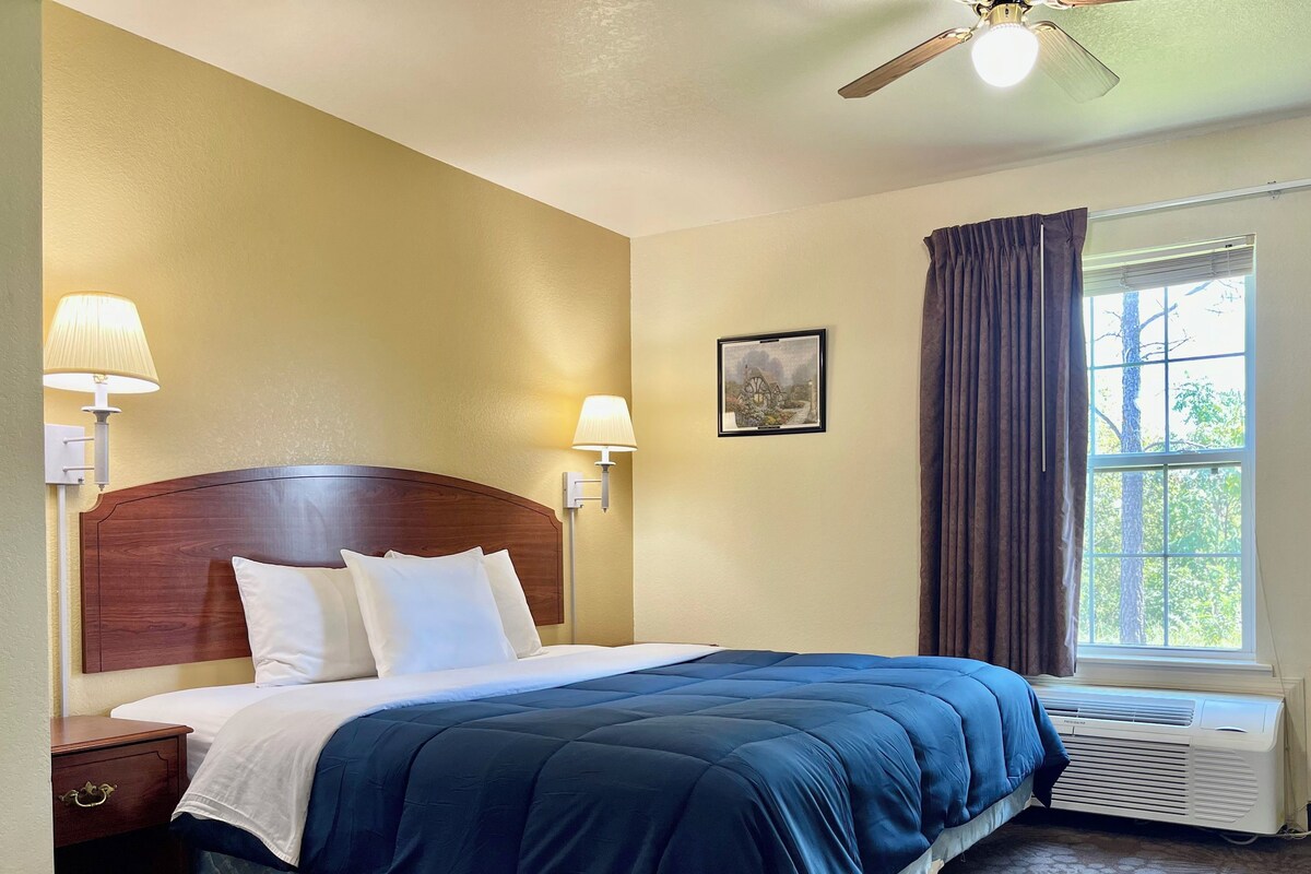A hotel room with a wooden headboard, a large bed with blue bedding, two bedside lamps, a window with curtains, and a wall-mounted picture.
