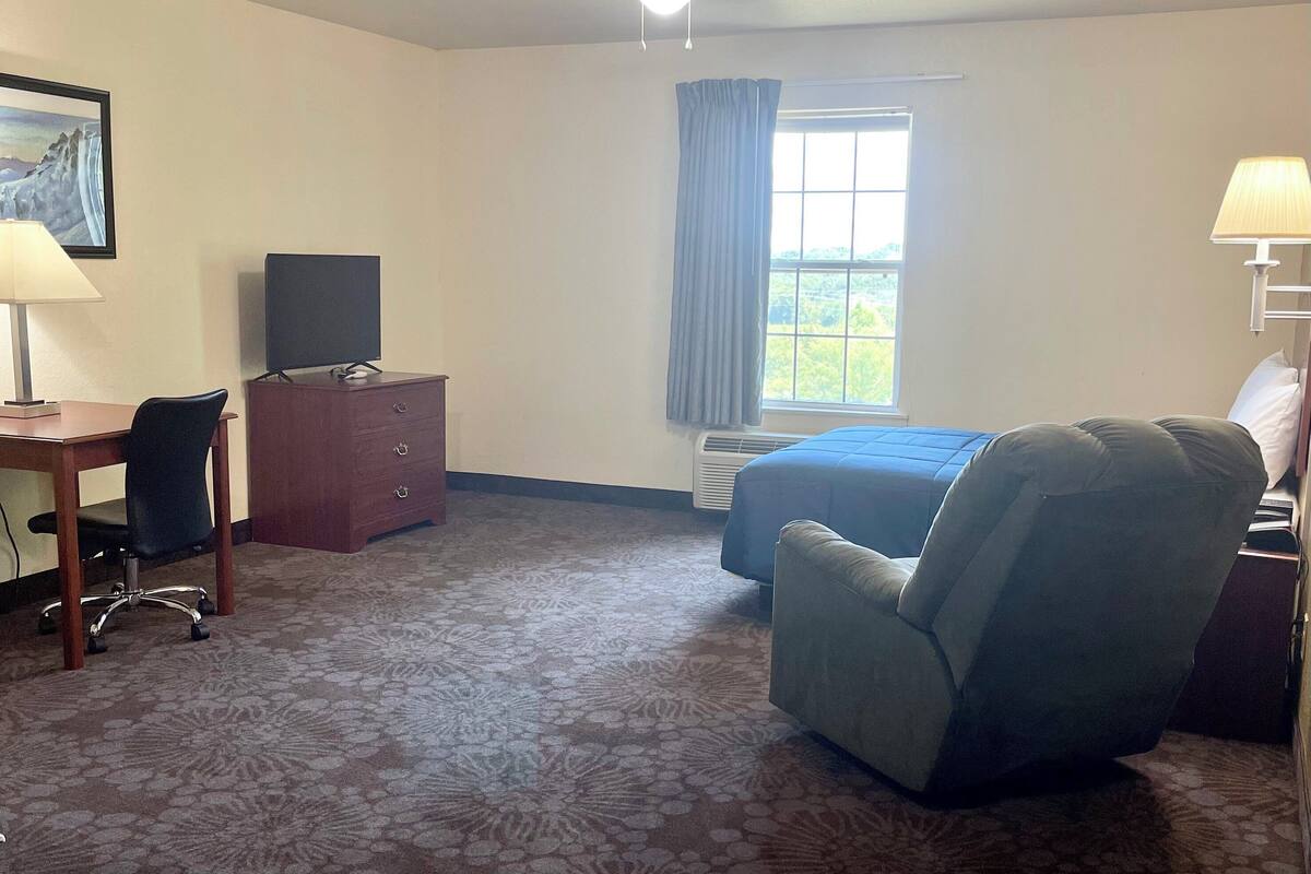 Ein Hotelzimmer mit Bett, Schreibtisch mit Stuhl, Fernseher, Deckenventilator, Fenster mit Vorhängen und Lampe.