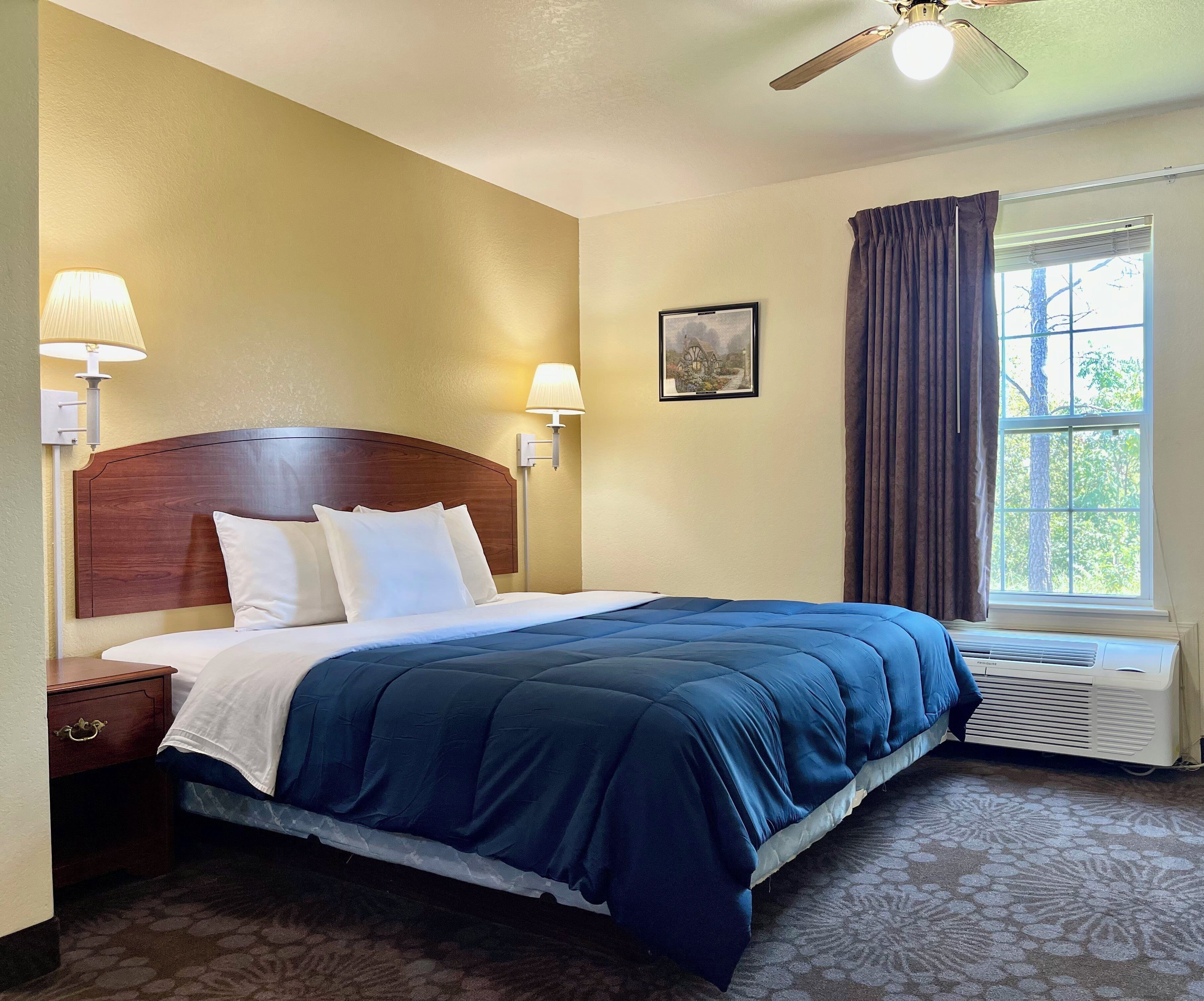 Une chambre d’hôtel avec une tête de lit en bois, un grand lit avec une literie bleue, deux lampes de chevet, une fenêtre avec des rideaux et un tableau accroché au mur.