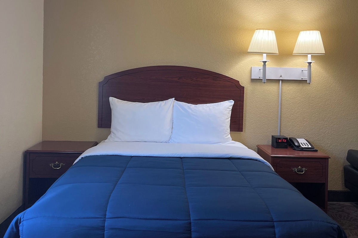 A hotel room with a bed, two bedside lamps, a nightstand with a phone, and a wall clock.