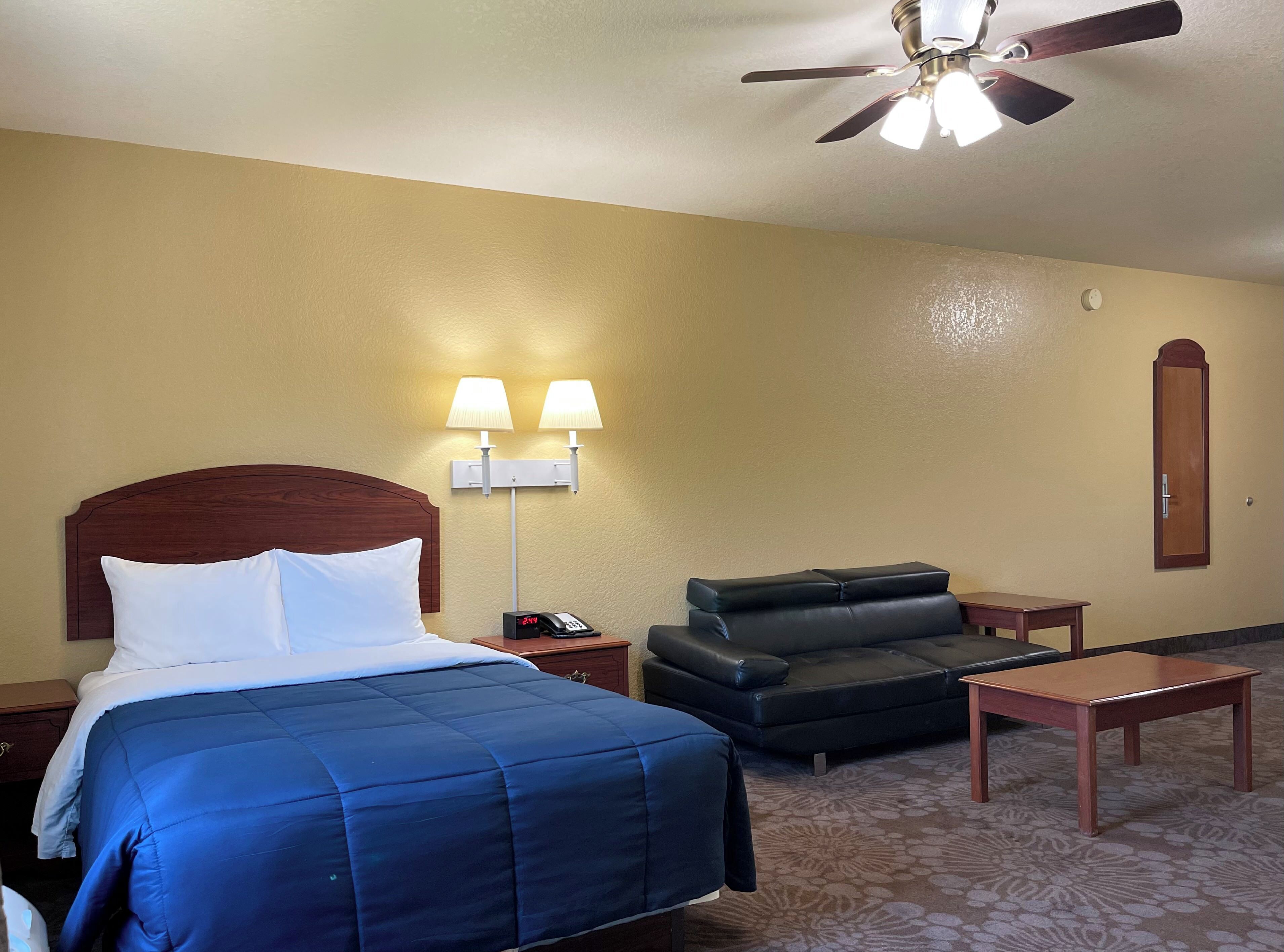 A hotel room with a bed, a desk, a chair, a nightstand, a lamp, a ceiling fan, and a wall clock.