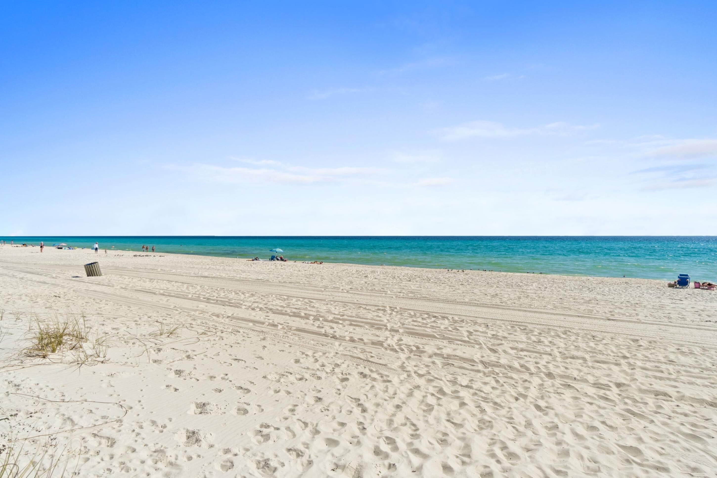 A wide sandy beach with footprints and a few people, extending to a calm sea under a clear sky.