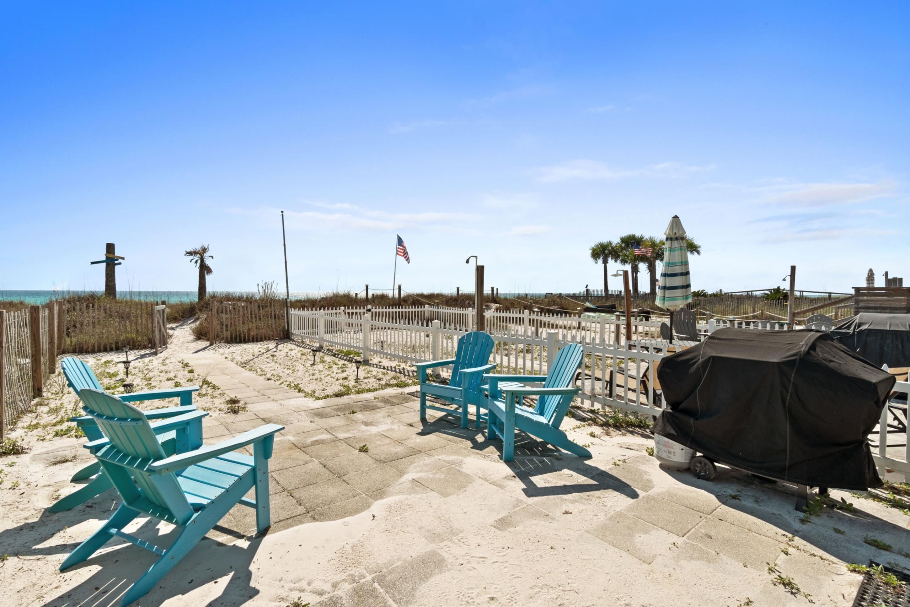 A beach patio with Adirondack chairs, a grill, and a fence.