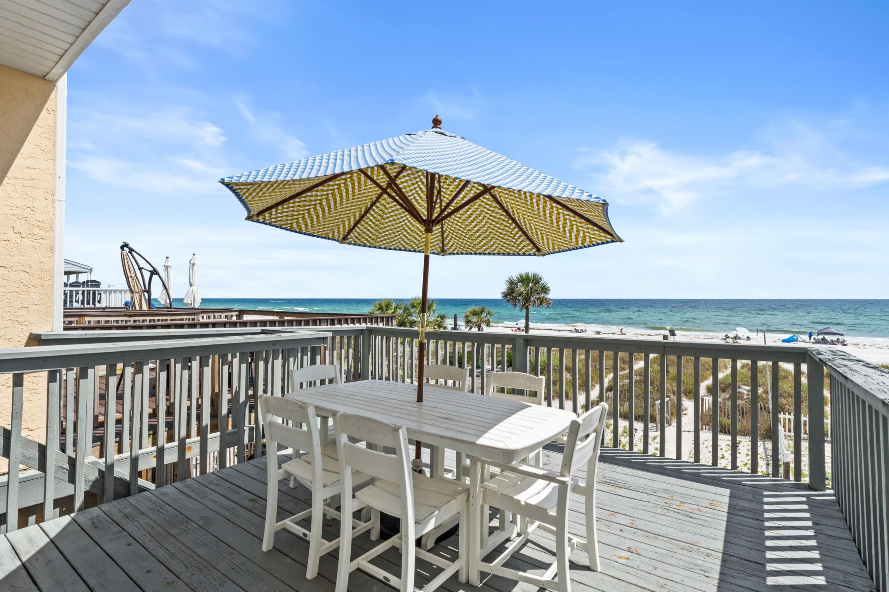 A wooden deck with a table and chairs, an umbrella, and a view of the beach and ocean.