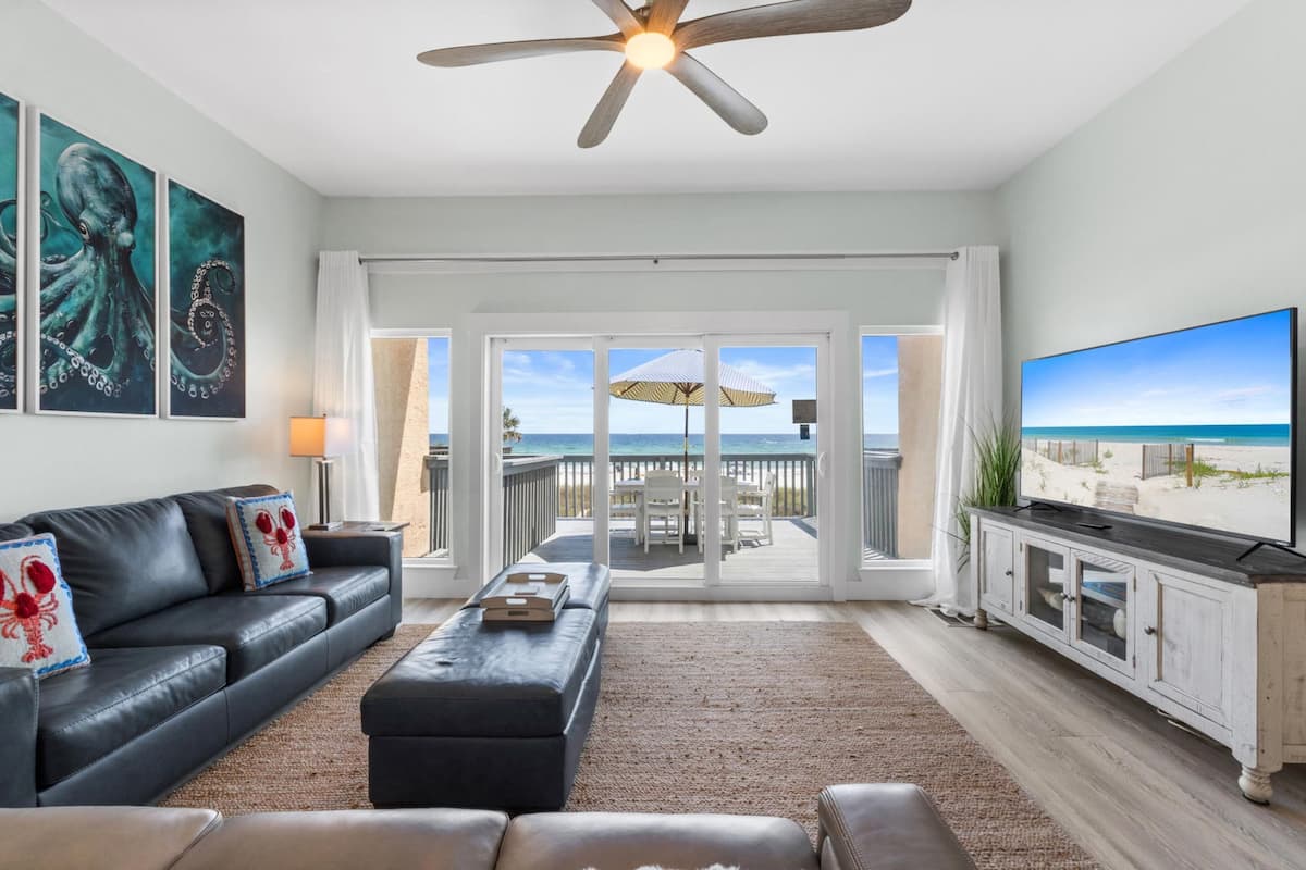 Ein Wohnzimmer mit großem Fernseher, Deckenventilator, einer Couch mit Zierkissen, einem Couchtisch und Blick auf den Strand durch Glastüren.