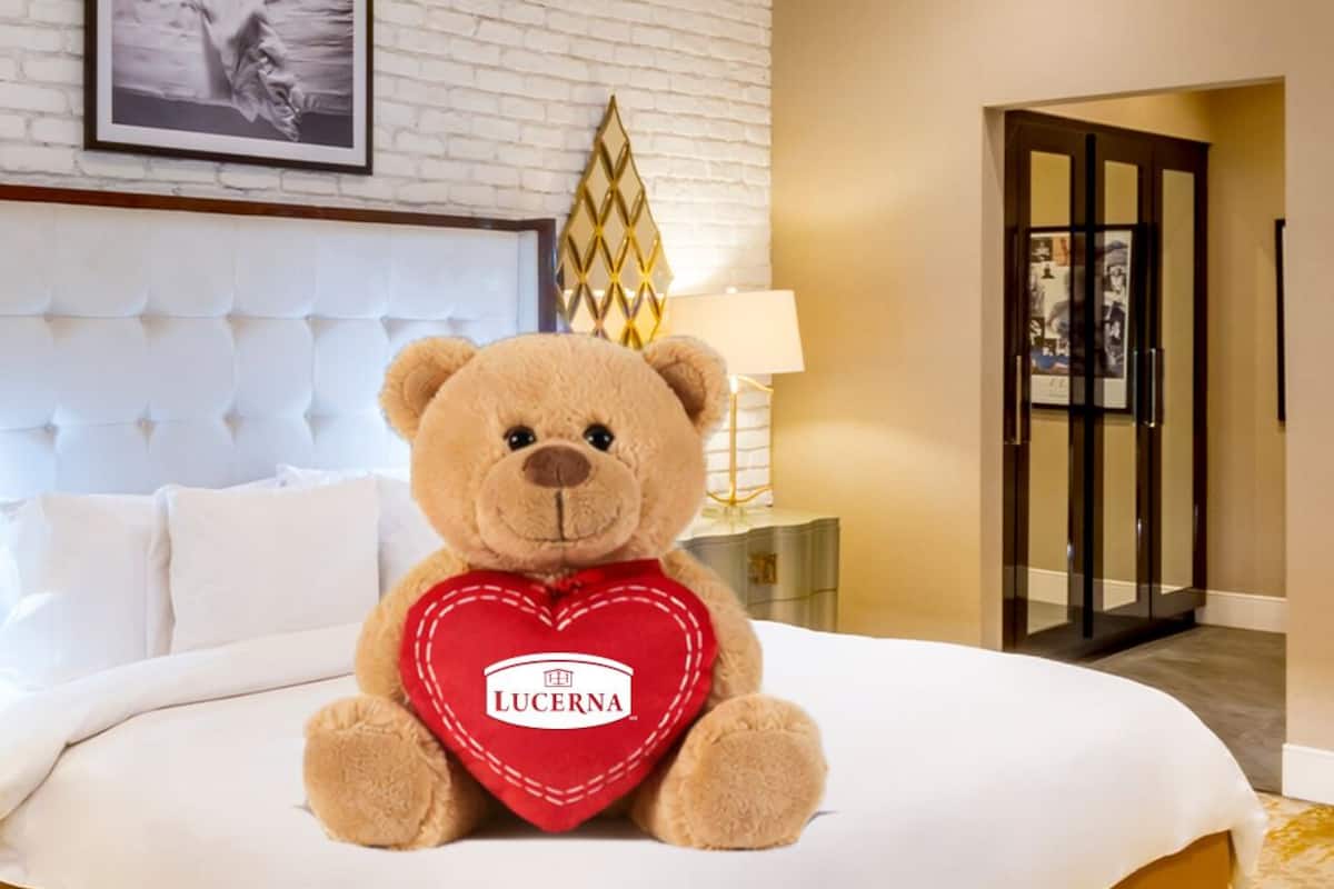 Een zachtaardig teddybeer houdt een hartvormig kussen vast, waarop "LUCERNA" staat, op een bed in een hotelkamer met een wit hoofdeinde, een lamp en een deur met ruitpanelen.