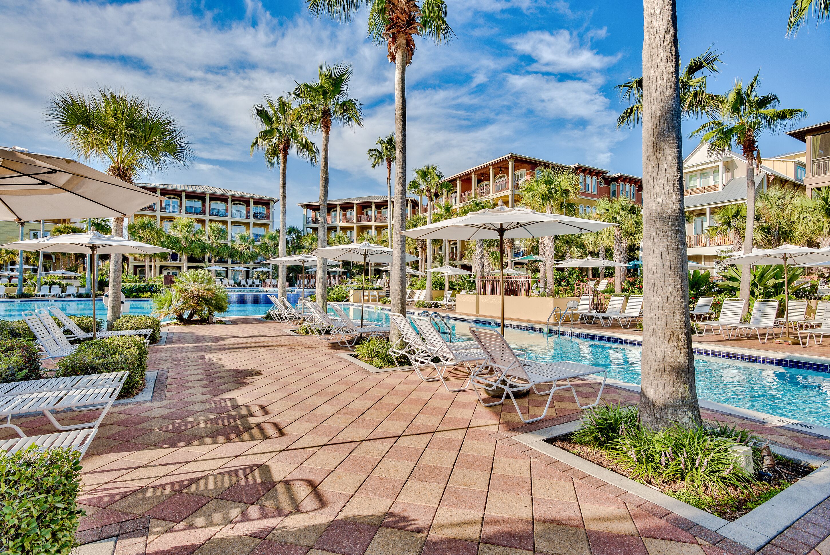 A resort pool area with lounge chairs and umbrellas, surrounded by palm trees and a building with balconies.