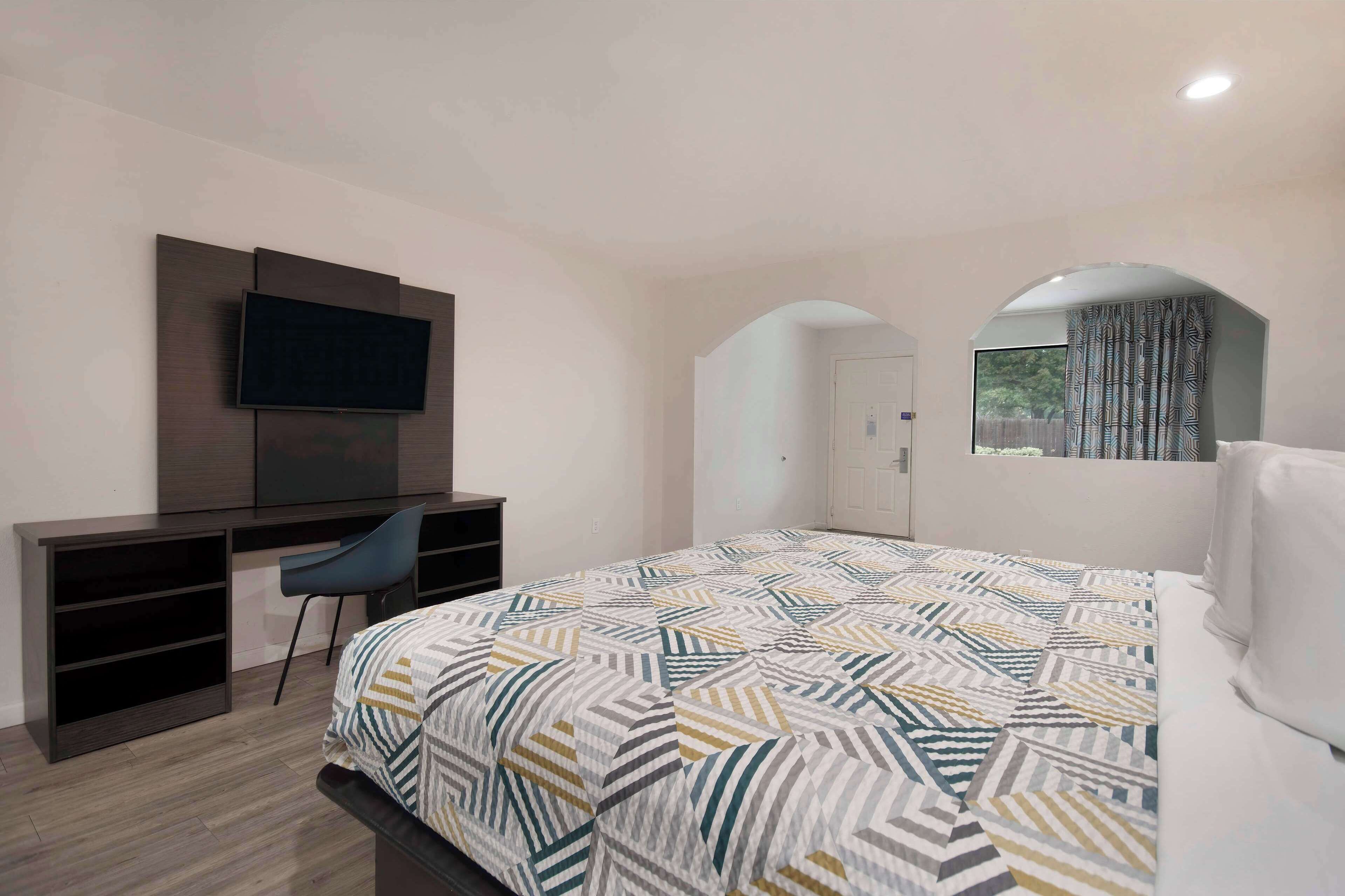 A bedroom with a bed, a desk, a chair, a TV, and a window with curtains.