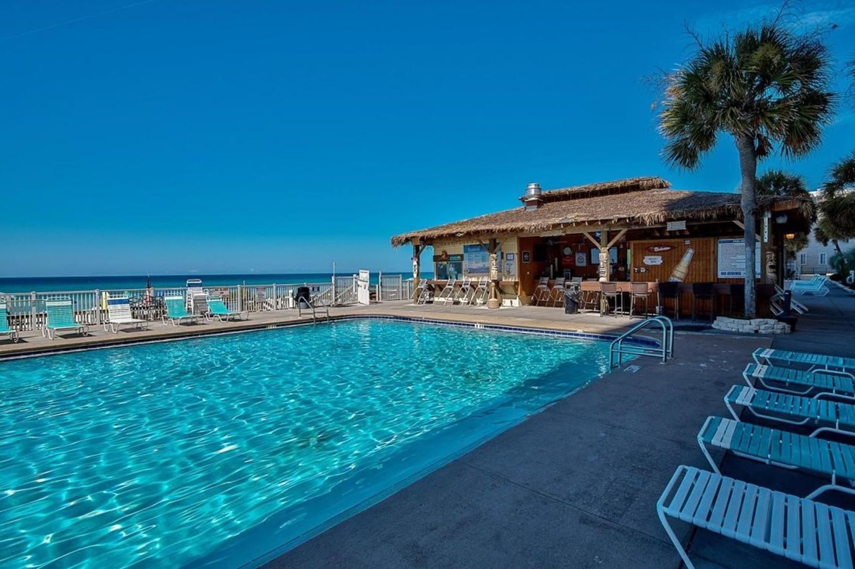 A poolside area with a clear view of the ocean, a bar with outdoor seating, and lounge chairs.