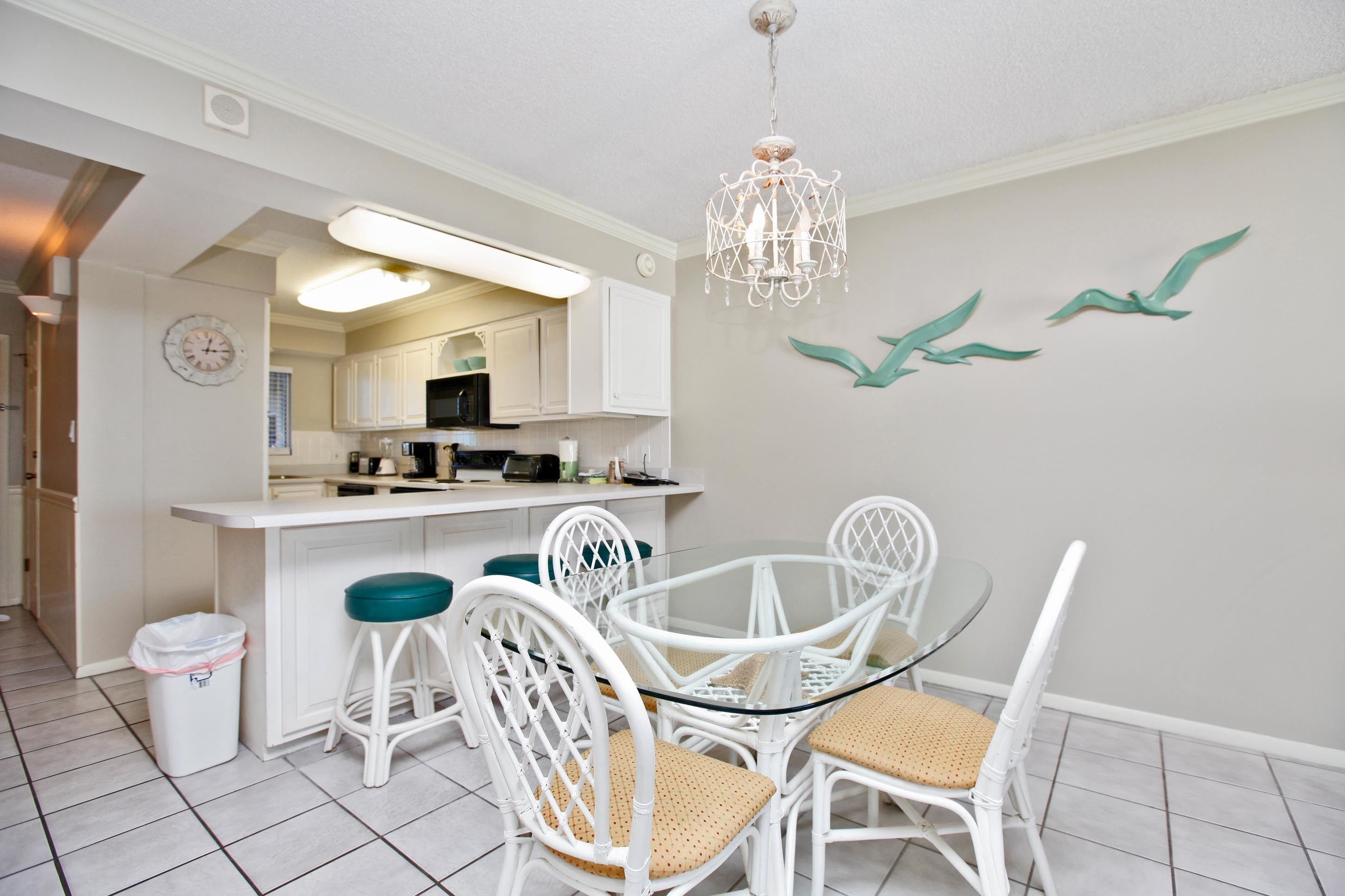 A modern kitchen with white cabinetry, a glass island, and a chandelier. There is a dining area with white chairs and a glass table.