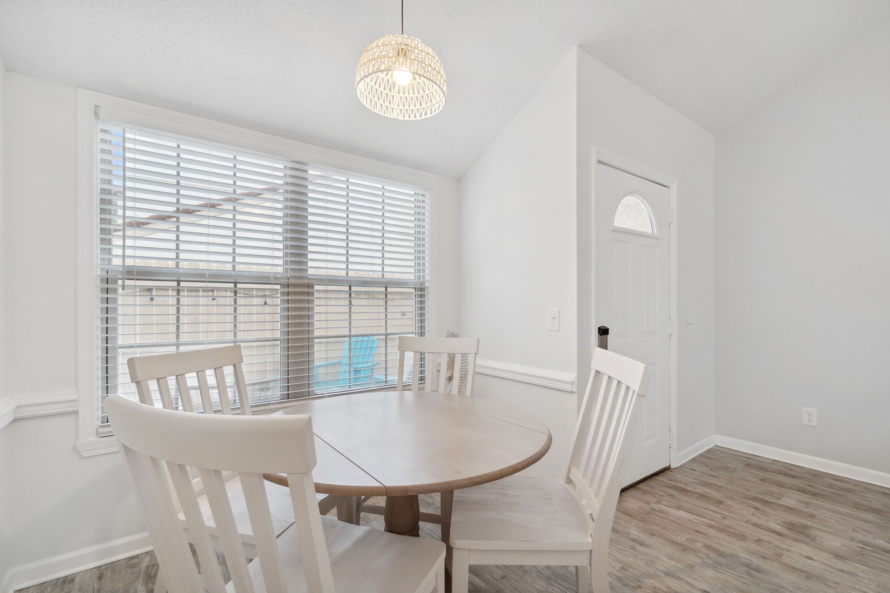 A dining area with a round table and four chairs, a hanging light fixture, and a window with blinds.