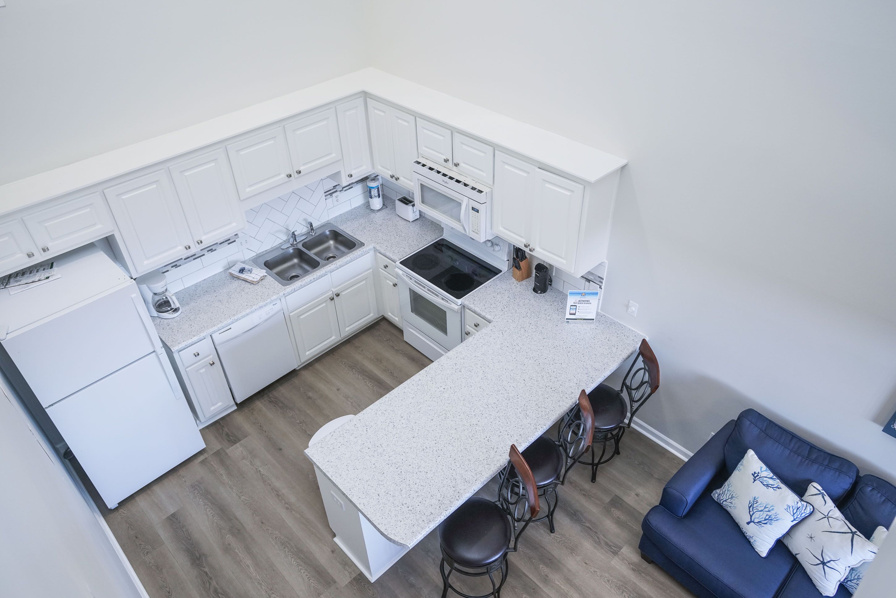 A modern kitchen with white cabinetry, a granite countertop, and a built-in oven. There are bar stools and a blue sofa with decorative pillows.