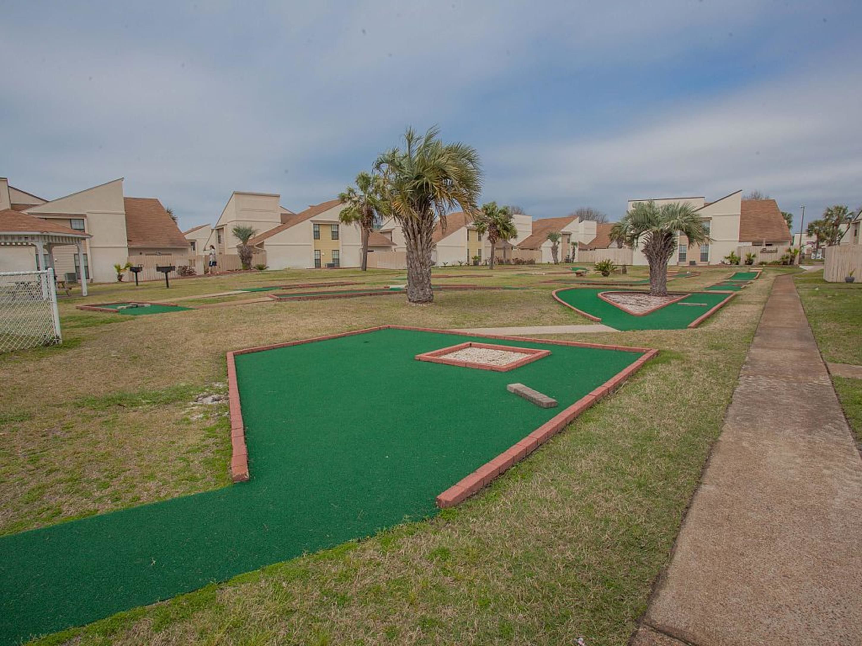 A mini-golf course with green artificial turf and red borders, surrounded by residential buildings with red roofs and beige walls.