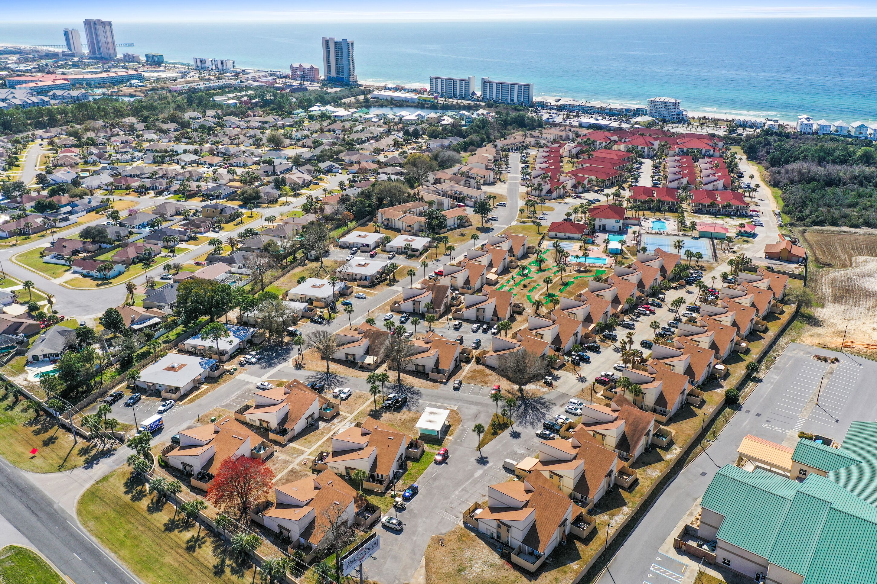 An aerial view of a residential area with houses, a parking lot, and a beach in the background.