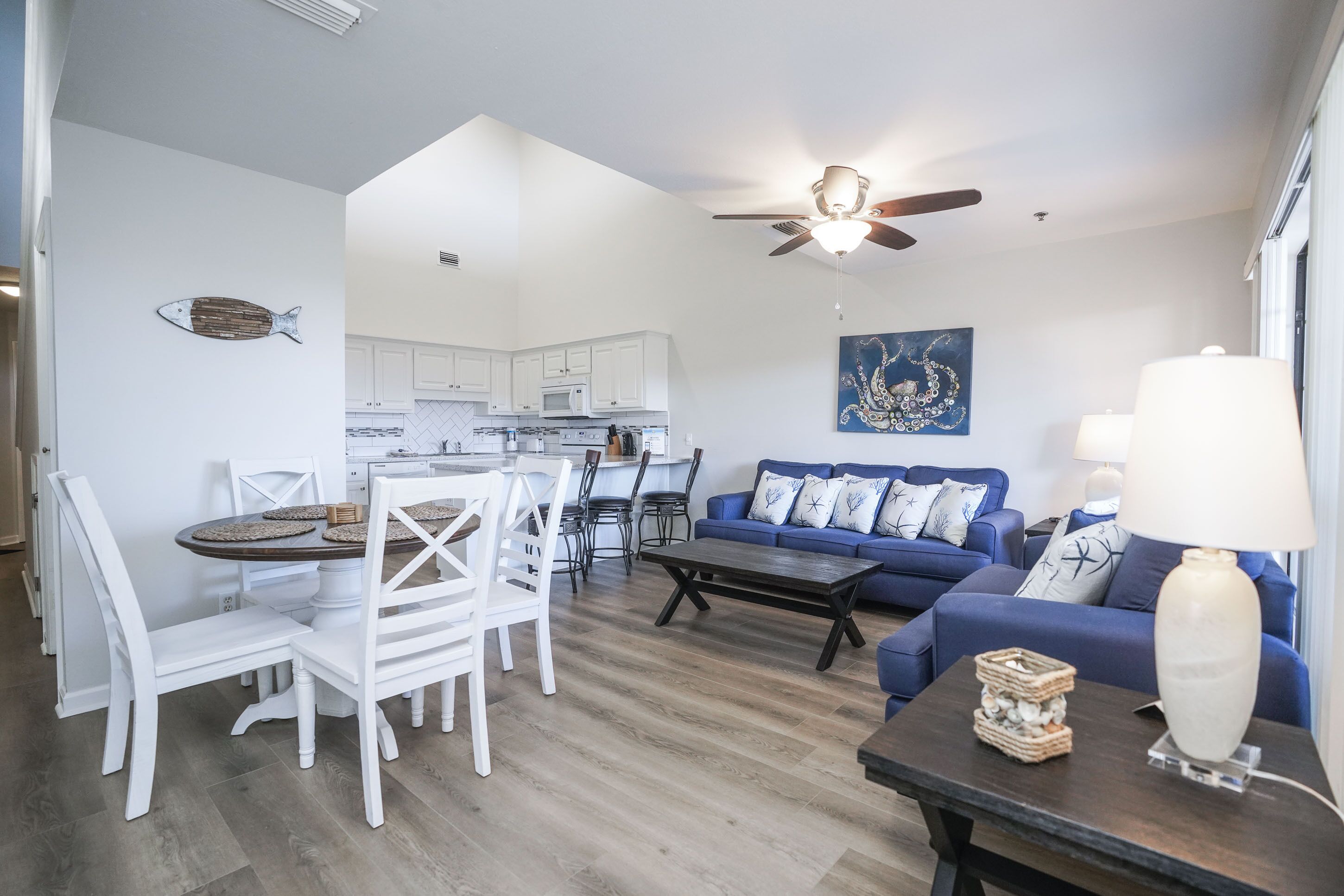 A modern living room with a dining area, a ceiling fan, a blue sofa, and a kitchen in the background.