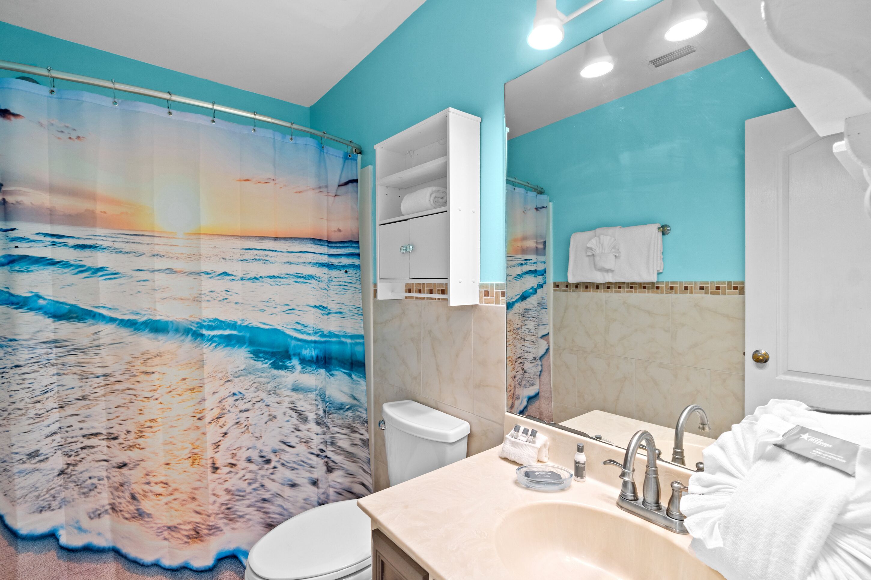 Une salle de bain avec un rideau de douche à motifs de plage, des toilettes blanches, un lavabo avec un miroir et un porte-serviettes.