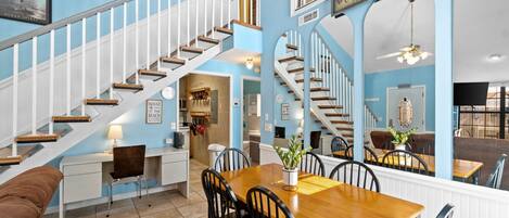 A spacious living area with a wooden dining table, black chairs, and a brown sofa. A staircase with white railings leads to another level.
