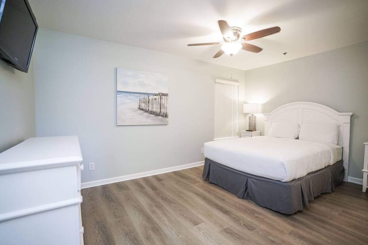 Een slaapkamer met een bed, een plafondventilator, een nachtkastje, een lamp, een dressoir en een schilderij van een pier.