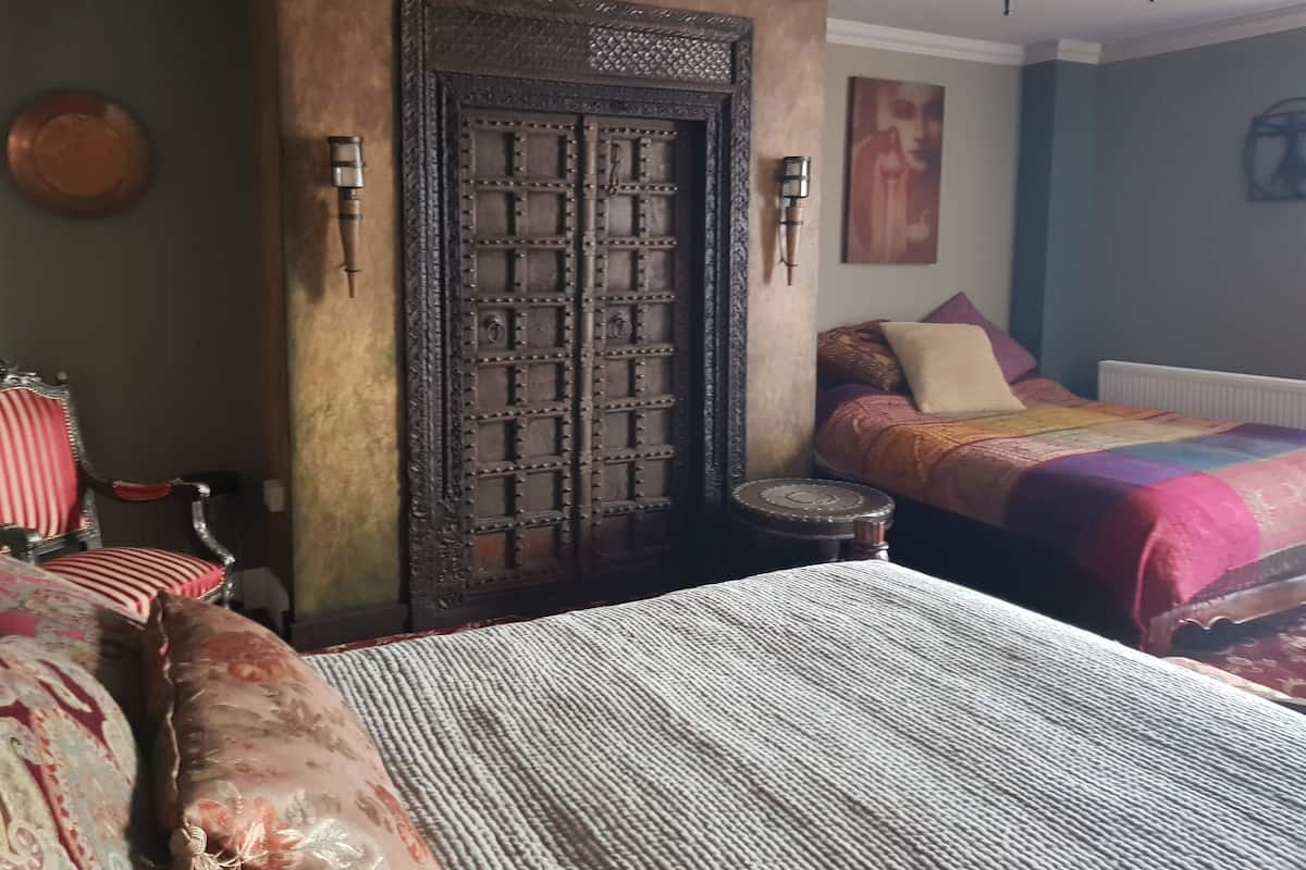 A bedroom with a large wooden door, a bed with patterned bedding, a small stool, and a chair with red cushions.