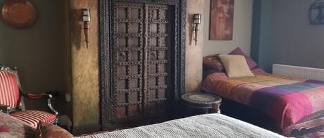Un dormitorio con una puerta de madera grande, una cama con ropa de cama estampada, un taburete pequeño y una silla con cojines rojos.