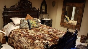 A bedroom with a large bed, a wooden headboard, a nightstand, a mirror, and a decorative wall piece. - The Covenstead (Glastonbury)