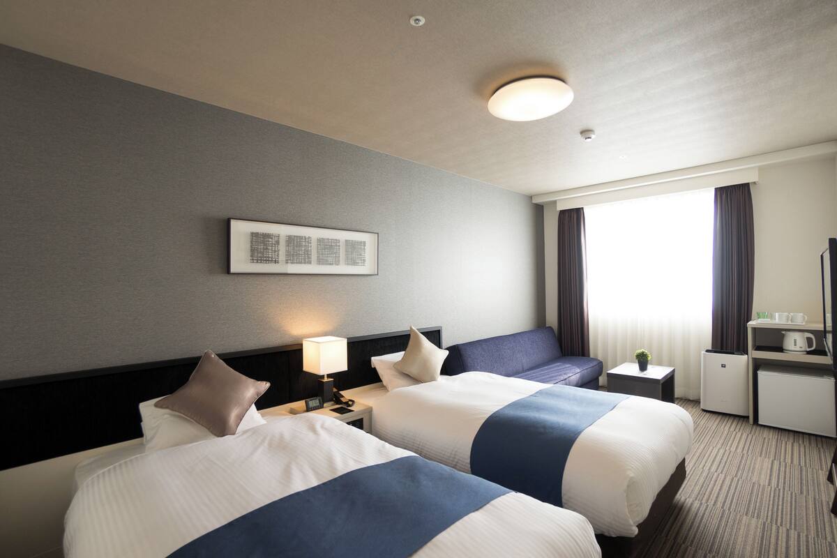 Een hotelkamer met twee bedden, een nachtkastje met een lamp, een kleintje tafeltje met een plant, een bureau met een koffiezetapparaat en een raam met gordijnen.
