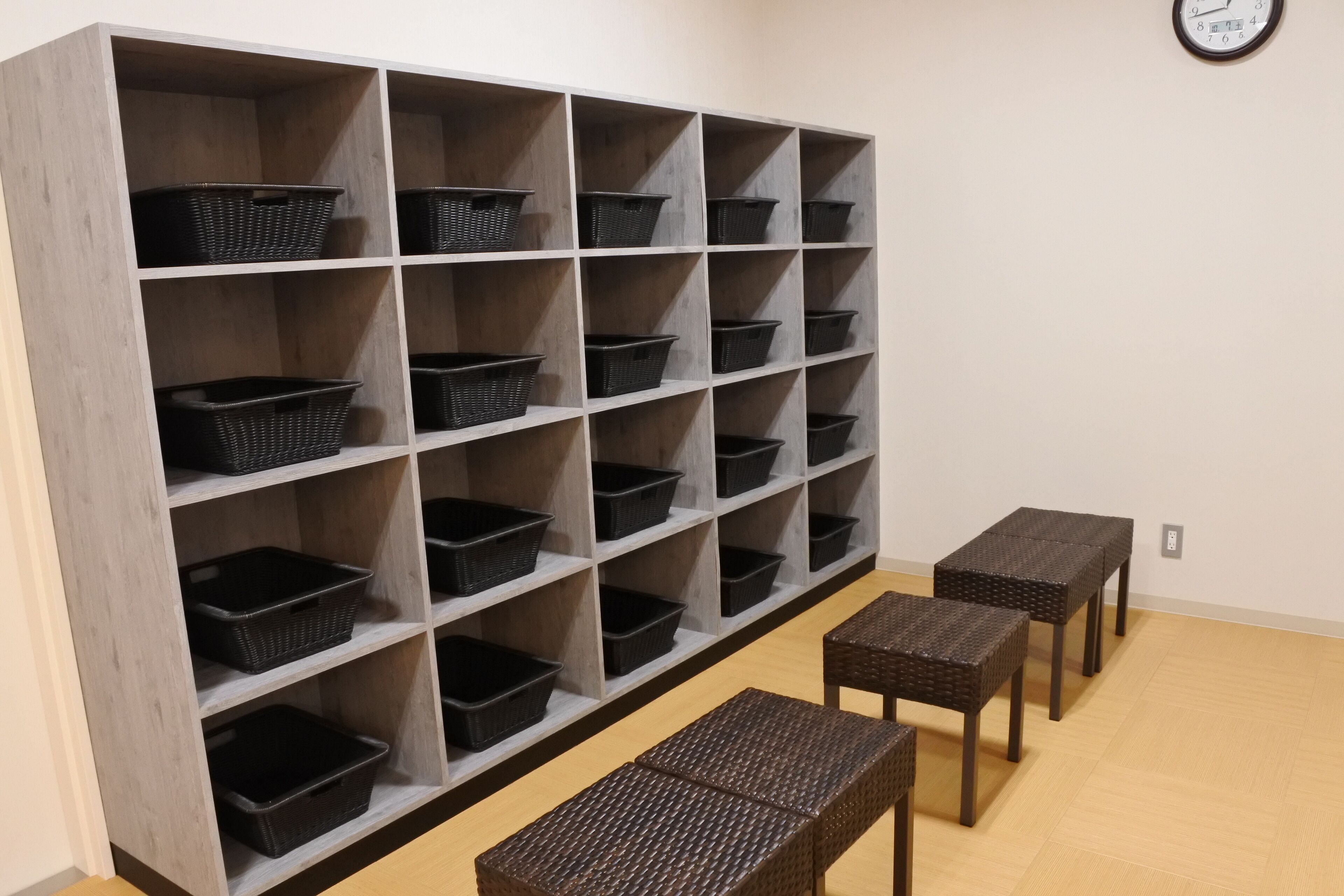 A room with a large shelving unit filled with black baskets, a clock on the wall, and three wicker tables.