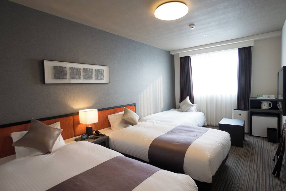Een hotelkamer met twee bedden, een bureau, een stoel, een lamp, een koffiezetapparaat en een raam met gordijnen.