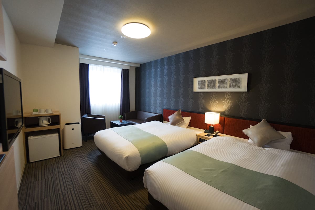 Een hotelkamer met twee bedden, een bureau, een stoel, een kleine koelkast en een raam met gordijnen.