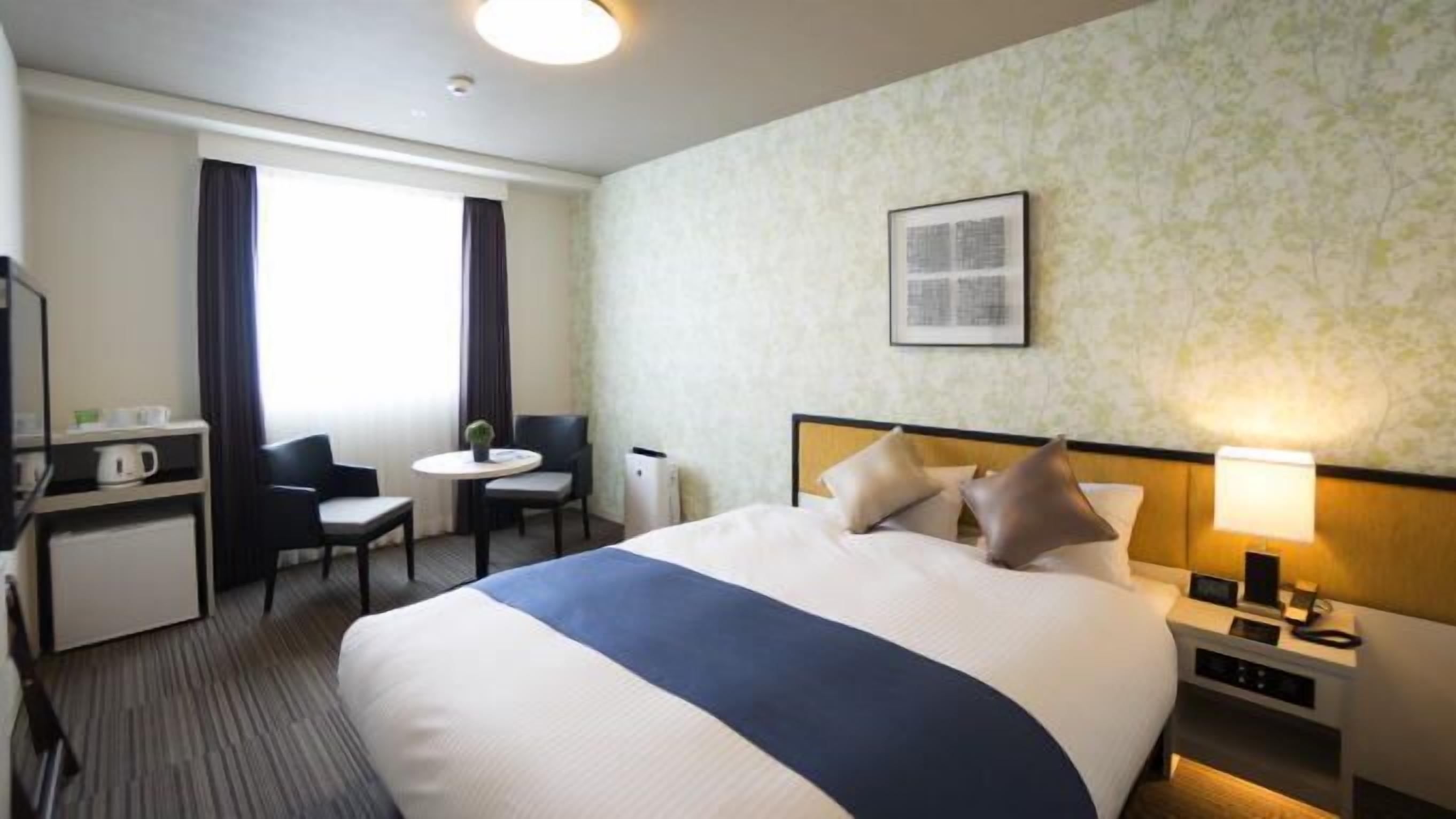 Une chambre d’hôtel avec un lit, un bureau, une chaise, une petite table, une lampe et un tableau au mur.