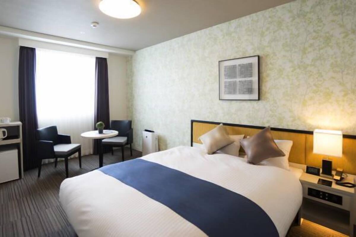 Een hotelkamer met een bed, een bureau, een stoel, een klein tafeltje, een lamp en een foto aan de muur.