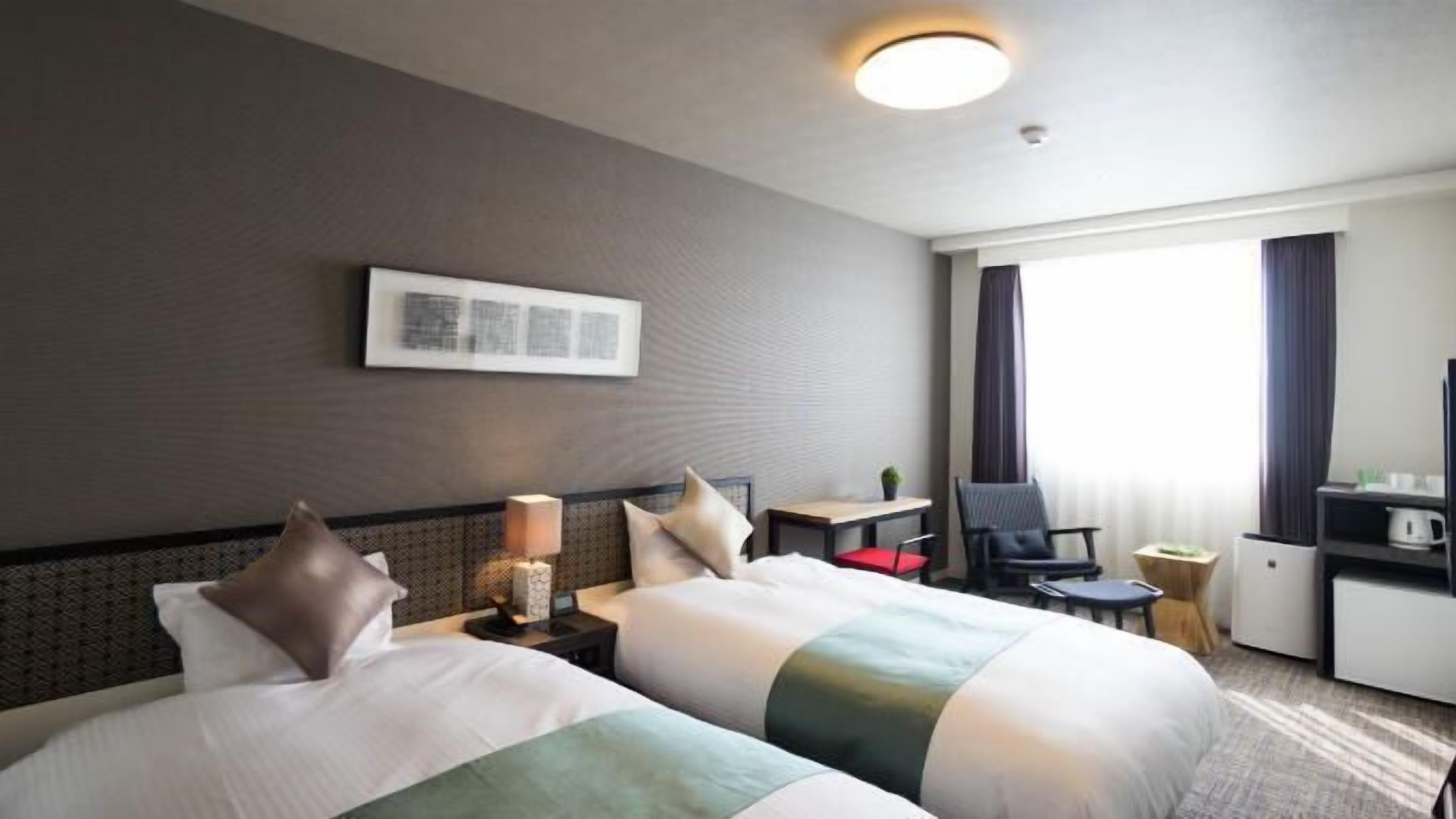 Une chambre d’hôtel avec deux lits, un bureau, une chaise, une lampe et une fenêtre avec des rideaux.