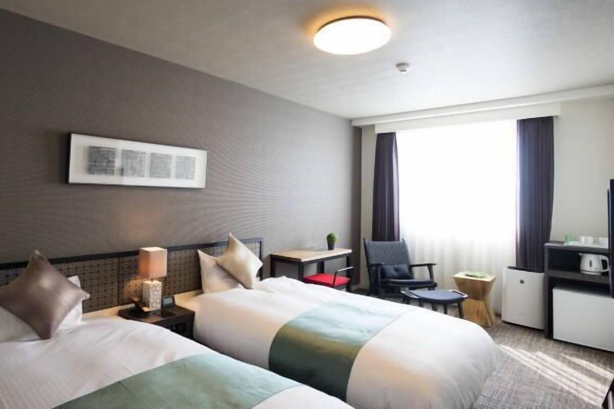 Een hotelkamer met twee bedden, een bureau, een stoel, een lamp en een raam met gordijnen.