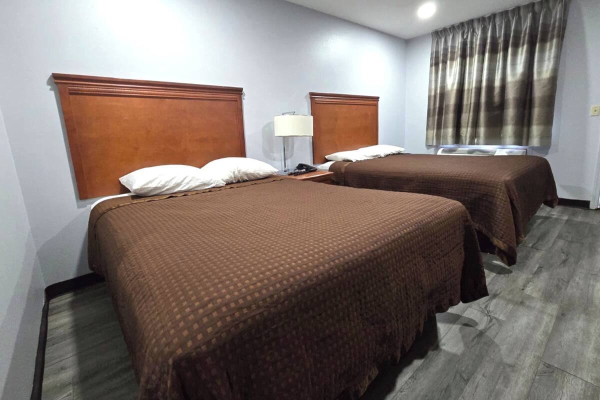A hotel room with two beds, a nightstand, a lamp, and a window with curtains.