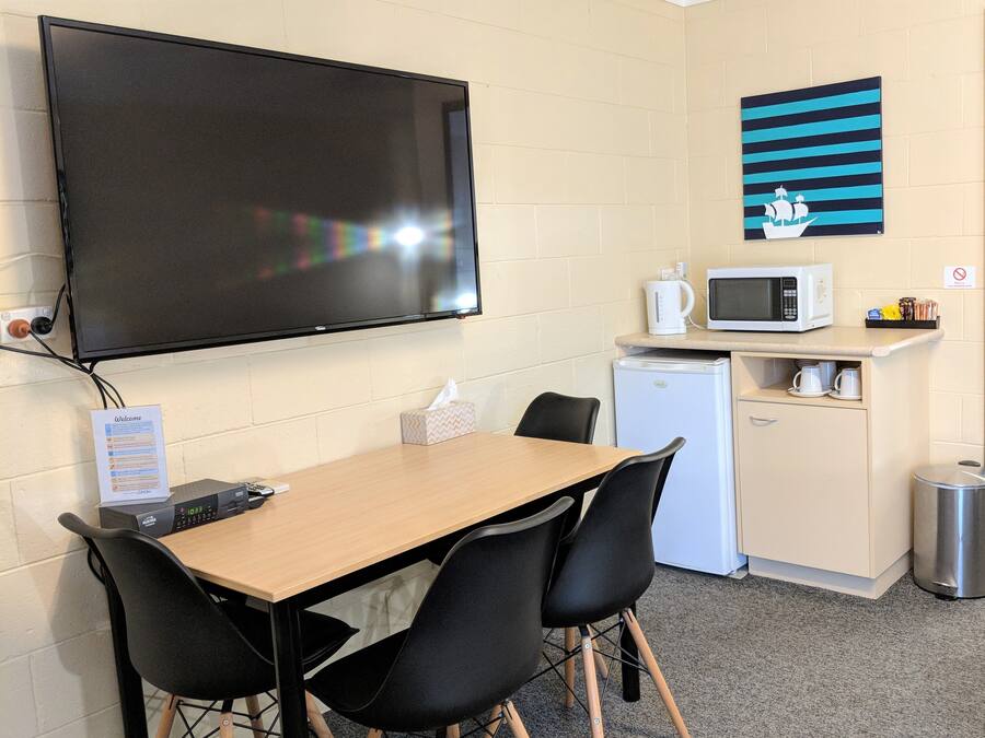 A small kitchenette with a microwave, kettle, and coffee maker. There is a TV mounted on the wall, a table with chairs, and a trash can.