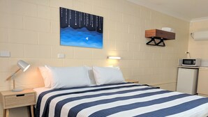 Habitación de hotel con cama, mesita de noche con lámpara, un cuadro en la pared, repisa con toalla y microondas.