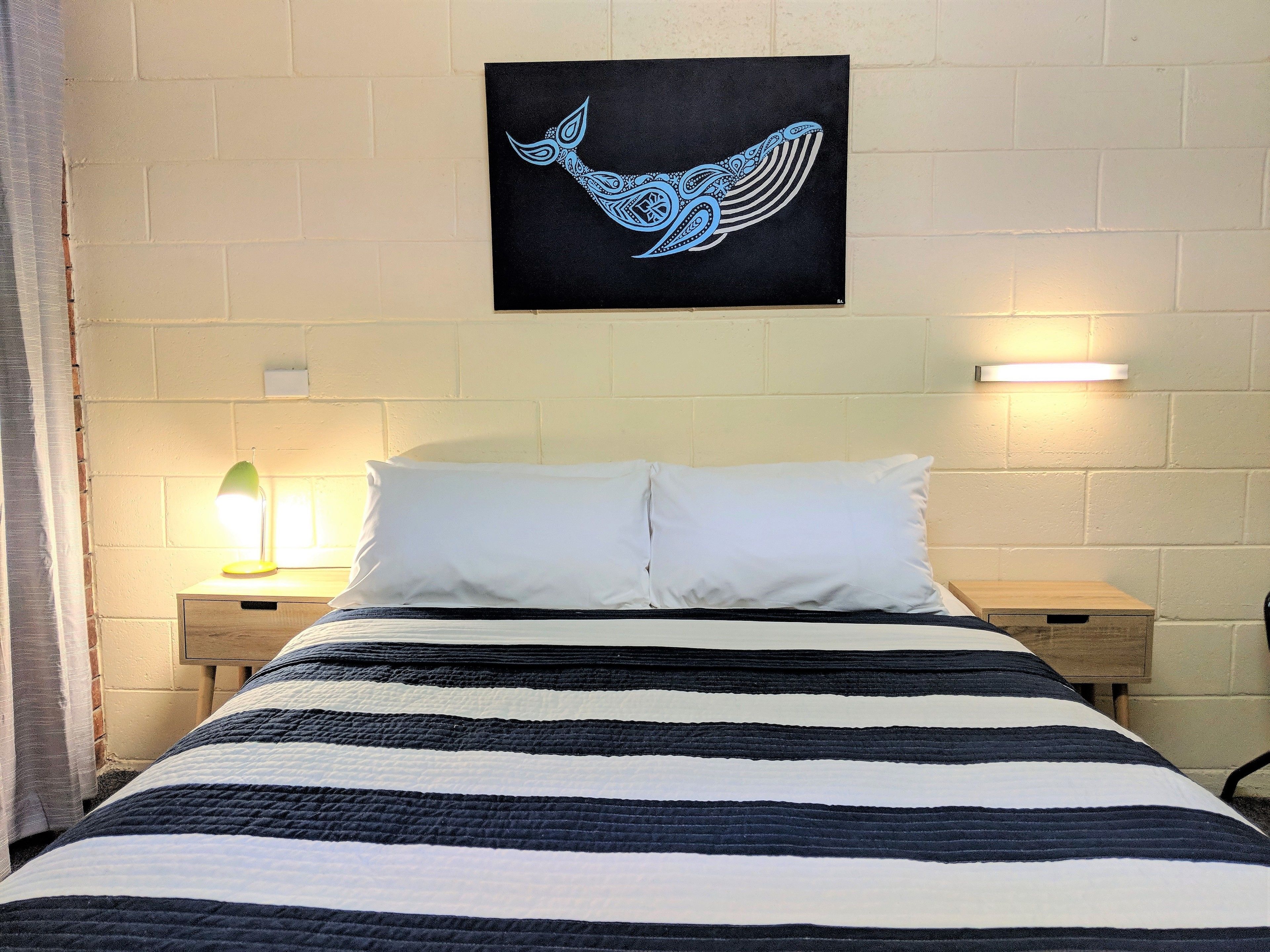 A bed with a striped bedspread, two white pillows, and a wall-mounted artwork of a blue whale. There are two bedside tables with lamps, and a white brick wall.