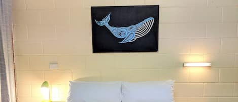 Una cama con colcha a rayas, dos almohadas blancas y una obra de arte en la pared de un tiburón ballena. Hay dos mesitas de noche con lámparas y una pared de ladrillo blanco.