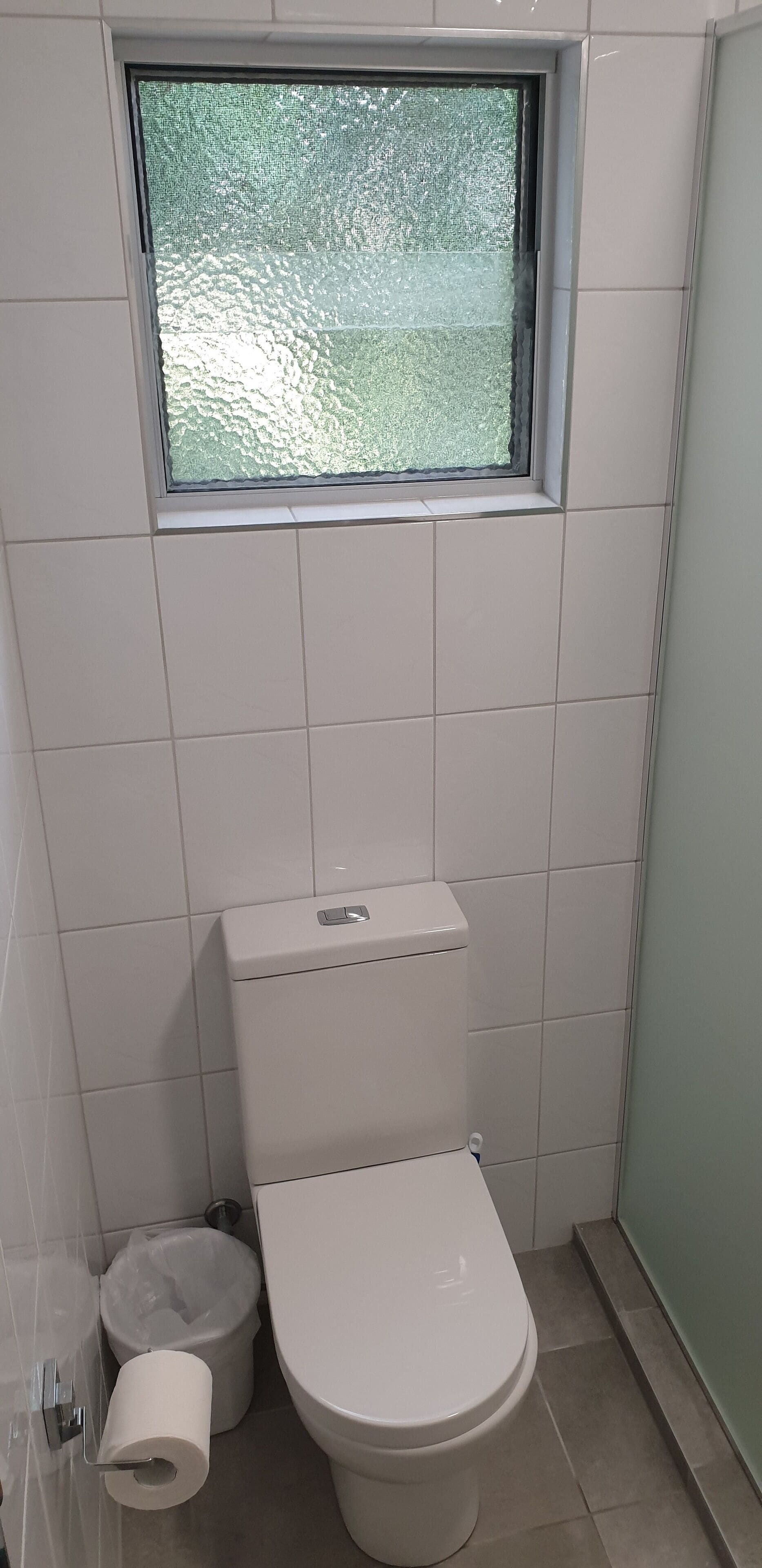 Un baño pequeño y limpio con inodoro, un cesto de basura y una ventana sobre el inodoro.